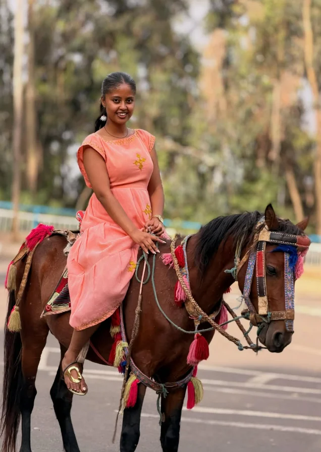 Efratah in a coral dress riding a decorated horse near Entoto